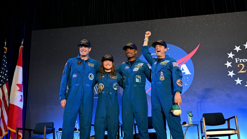 CSA (Canadian Space Agency) astronaut Jeremy Hansen and NASA astronauts Reid Wiseman, Victor Glover, and Christina Koch smile at the crowd during a news conference. They are all wearing blue jumpsuits with patches on the arms and chest areas. Wiseman raises his right fist in a victorious pose.