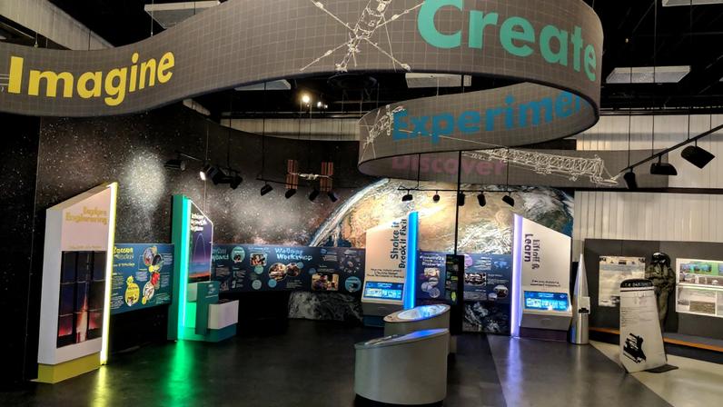 A gray banner with the words Imagine, Create, Experiment and Discover hangs above a colorful exhibit with panels with information about engineering and sounding rockets