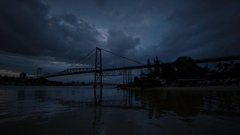 Vista aérea de Florianópolis ao entardecer, com a Ponte Hercílio Luz em destaque e céu alaranjado refletindo no mar.