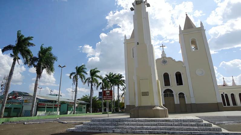 Praça central de Monsenhor Tabosa CE com igreja ao fundo, céu azul e vegetação de praça