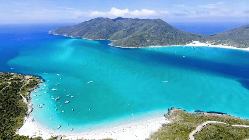 Vista aérea da orla de Arraial do Cabo RJ, com mar azul-turquesa, barcos e faixa de areia clara