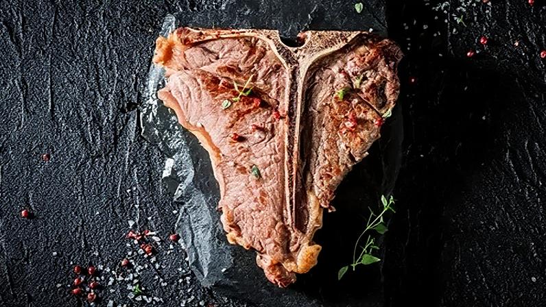 A thick T-bone steak sits against a black background, scattered with red peppercorns, rock salt and herb (Credit: Alamy)
