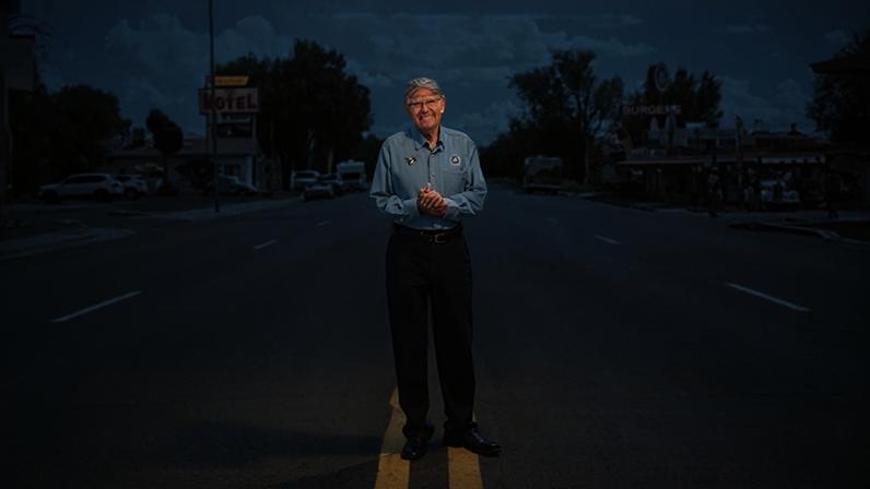 Johnny Kompar Angel Delgadillo stands in the middle of wide street on Route 66 in a postcard-style image reading "Greetings from Route 66"