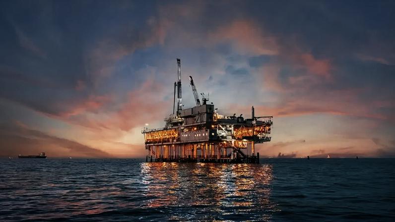 Imagem de uma plataforma de petróleo no mar ao pôr do sol, com céu colorido de tons de rosa e laranja, refletindo na água.