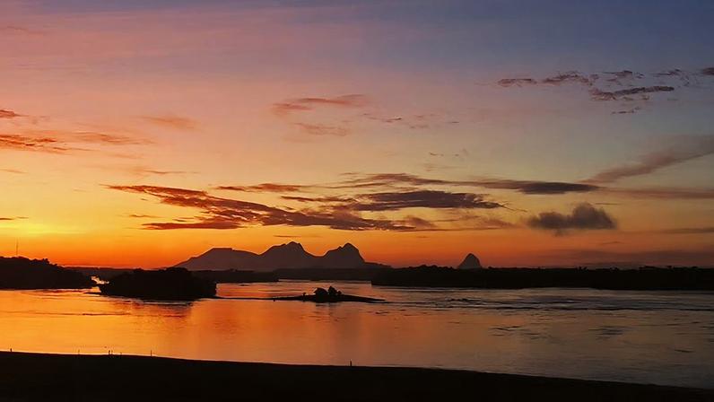 Pôr do sol refletido no Rio Negro, com vegetação amazônica e cadeias de montanhas ao fundo.