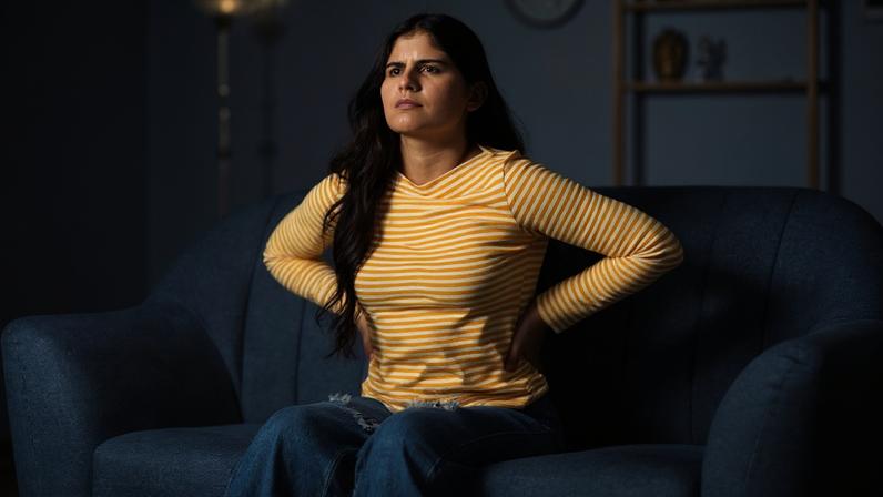 Foto colorida de mulher sentada em sofá e com as mãos na lombar - Metrópoles