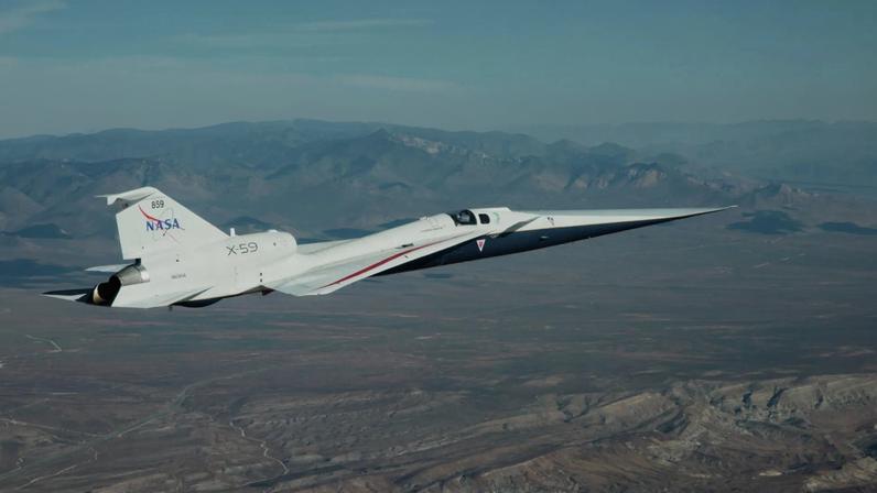 NASA’s X-59 flies above the Mojave Desert on a clear day. The white aircraft has light gray, red, and blue accents, with a NASA logo and the number 859 on its tail. It appears flying level over the desert landscape, with a mountain range visible on the horizon and a trail of clouds above.