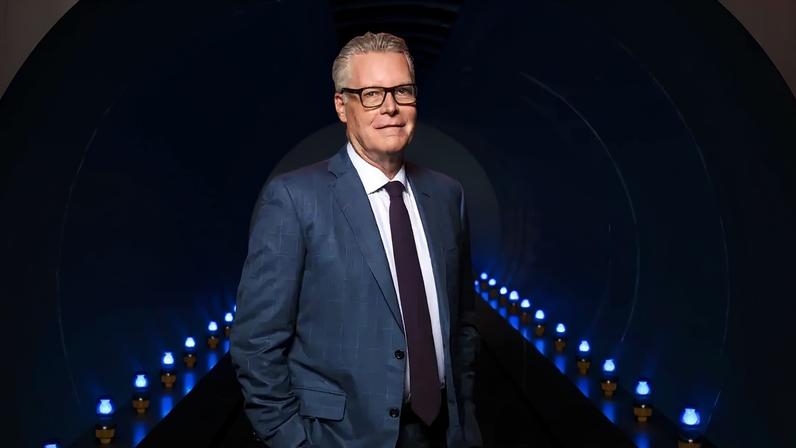 Ed Bastian stands for a photograph. He is wearing a suit and tie and has his hands in his pockets. There are rows of blue lights on either side of him.