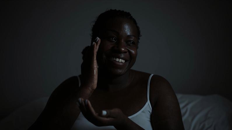 Imagem mostra mulher negra, com camiseta branca, sorrindo e passando creme no rosto - pele saudável precisa de nutrientes