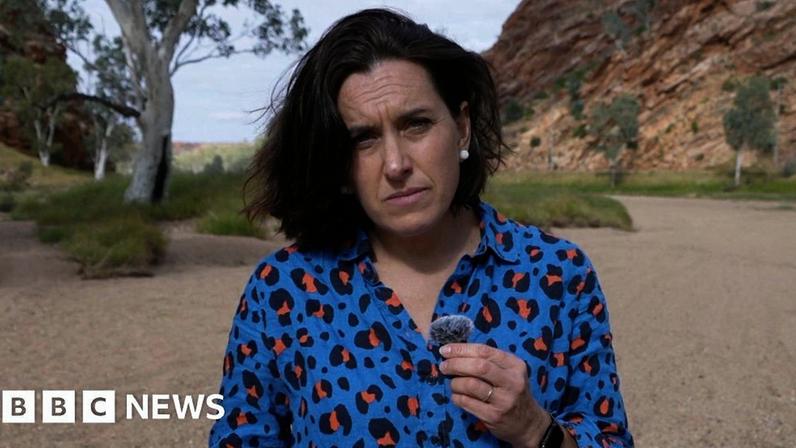 The BBC's Katy Watson wearing a blue leopard print blouse stands in front of a rocky outcrop