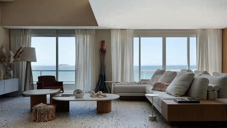 SALA DE ESTAR | Com vista do mar através das grandes aberturas, o ambiente é repleto design brasileiro, a exemplo do sofá Bass e da poltrona Isa, ambos de Jader Almeida. Mesas de centro Drop da Saccaro. Os tons de areia predominam nas paredes com textura Ornamentali Bege Bahia, da Diore Revestimentos, e no tapete de lã da Oriente-se. Entre as cortinas de linho da JRJ, com execução de Alex Fernandes Cortinas, está a escultura Entrelaço, da coleção colaborativa O Mar entre Nós de Juliana Pippi e estúdio BÁ. Luminária coluna de piso Realejo da Bertolucci, na Ouse Iluminação