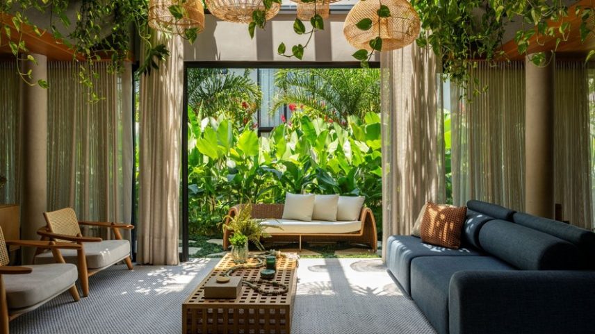 SALA DE ESTAR | A estética natural impera no ambiente, com luminárias no teto em fibra natural da Biasá Home ao lado de plantas pendentes, como jiboia e mini xanadu, além de maranta-charuto no corredor avarandado ligado pelo grande vão com cortinas da Anas Home Decor. O mobiliário - adquirido na Mobilare - mistura tons neutros com toques de azul e verde