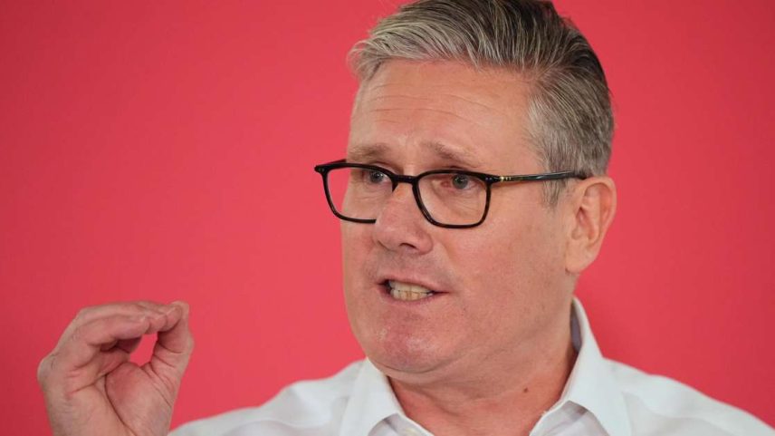 Primeiro-ministro britânico usando óculos com uma armação preta e uma camisa branca. Ele gesticula enquanto fala