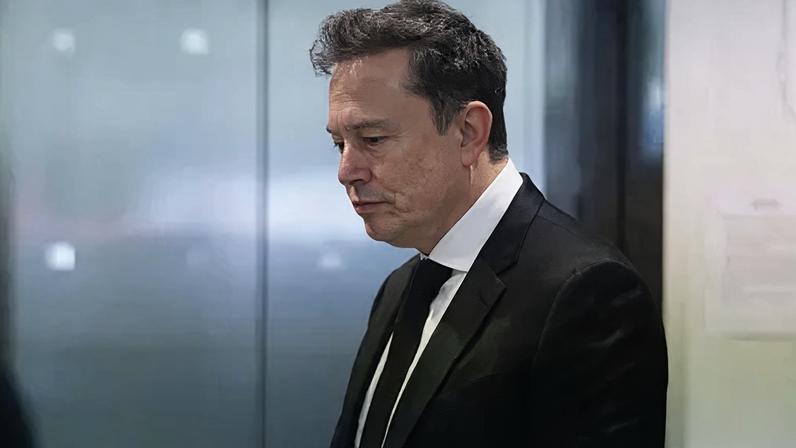 Musk, wearing a black suit, white shirt and black tie, looks down toward the floor while waiting to pass through security at a courthouse in California.