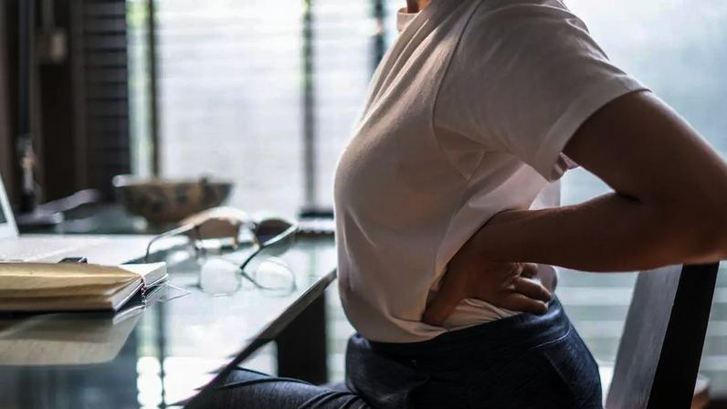 Uma mulher sentada à mesa coloca as mãos atrás das costas em sinal de dor