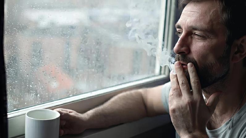 Homem tomando café e fumando um cigarro ao lado de uma janela.