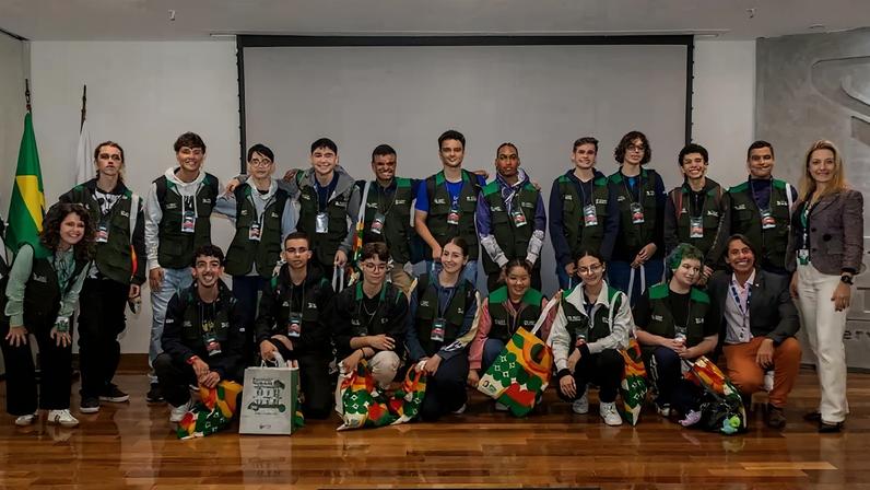 Grupo de jovens e adultos sorrindo, a maioria vestindo coletes verdes, posando em um palco de madeira com bandeiras e um telão branco ao fundo