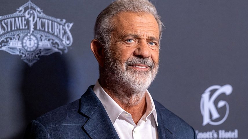 Mel Gibson, homem branco de cabelos grisalhos e barba, sorrindo levemente, vestindo blazer xadrez azul e camisa branca, em frente a um fundo escuro com logotipos