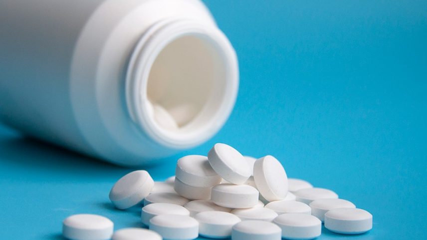 Frasco branco de remédio tombado, com várias pílulas brancas redondas espalhadas sobre uma superfície azul