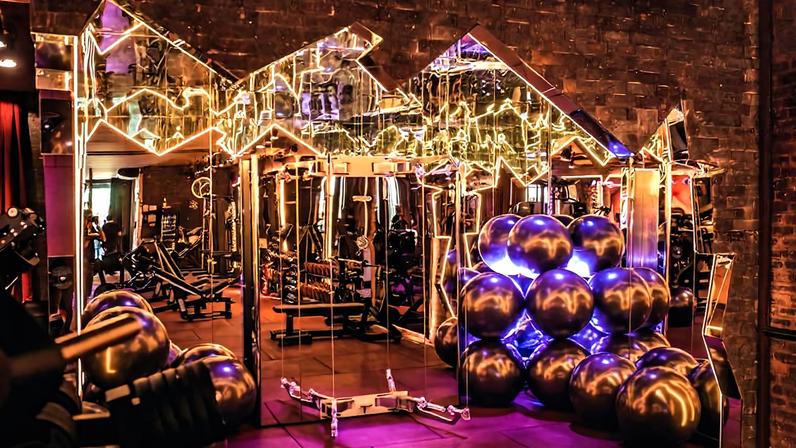 Imagem mostra uma sala com bolas de pilates cinzas, mas essa sala tem um aspecto de discoteca, um pouco escuro e com luzes de neon brancas em formatos geométricos