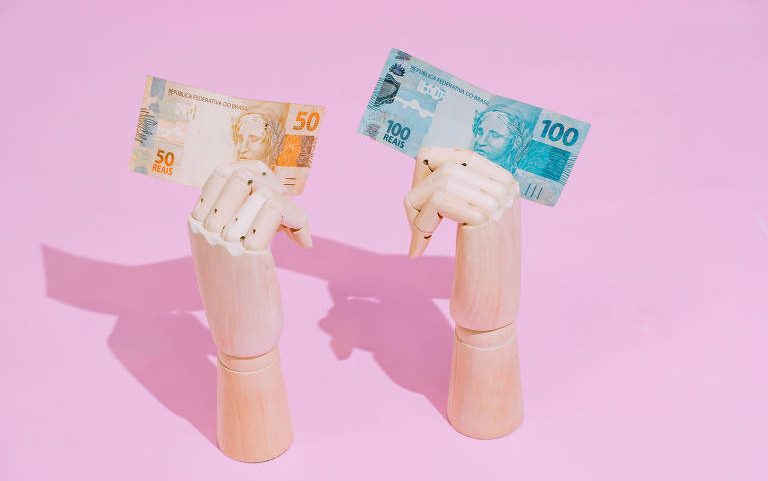 A imagem mostra duas mãos de madeira, uma segurando uma nota de 50 reais e a outra segurando uma nota de 100 reais. O fundo é rosa, criando um contraste com as notas e as mãos.