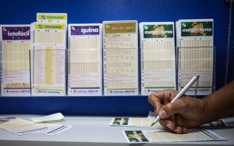Mão segurando caneta preenchendo bilhete da Mega-Sena sobre balcão branco. Ao fundo, seis cartazes de diferentes jogos de loteria, incluindo Lotofácil, Timemania, Quina, Dia de Sorte e Mega-Sena, fixados em parede azul.