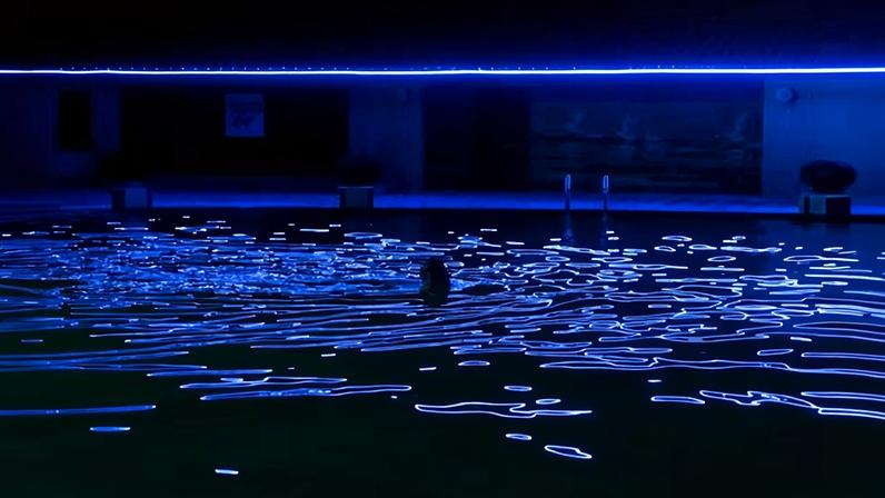 A person swims in a dark pool in Pyongyang with glowing blue light patterns projected on the surface