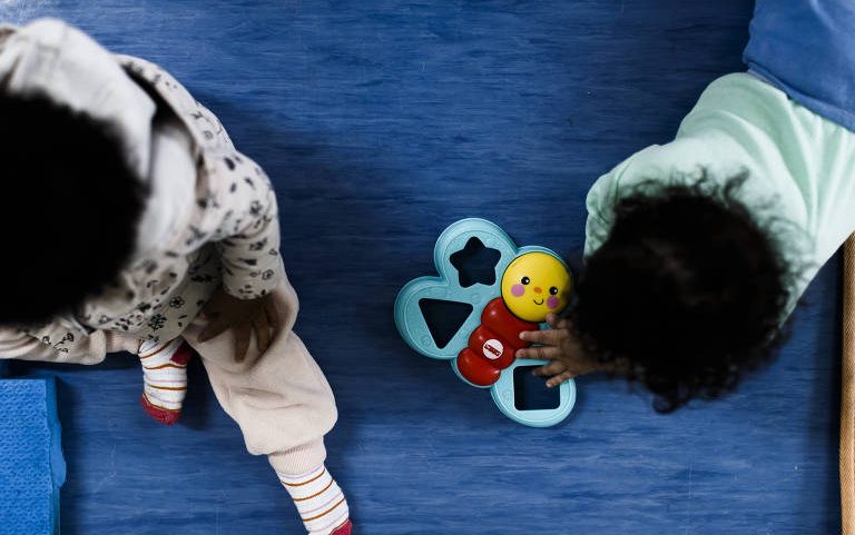 Duas crianças pequenas estão sentadas no chão azul. Uma delas, à esquerda, veste roupa clara com meias listradas, e a outra, à direita, usa camiseta azul clara. Elas interagem com um brinquedo de encaixe colorido em forma de lagarta com peças geométricas.