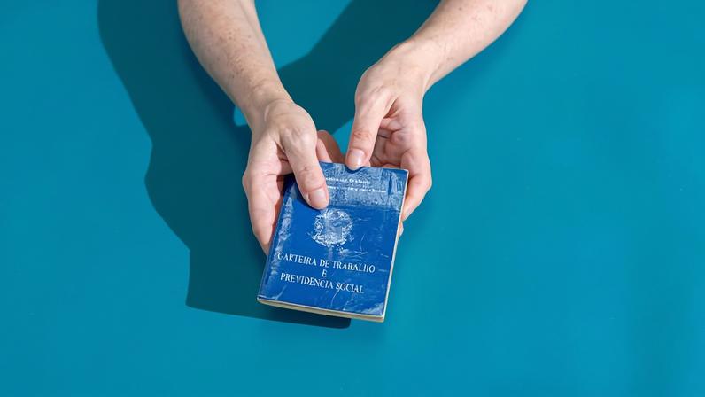 Duas mãos seguram um passaporte brasileiro azul contra um fundo azul liso. A luz cria sombra das mãos e do passaporte à esquerda.