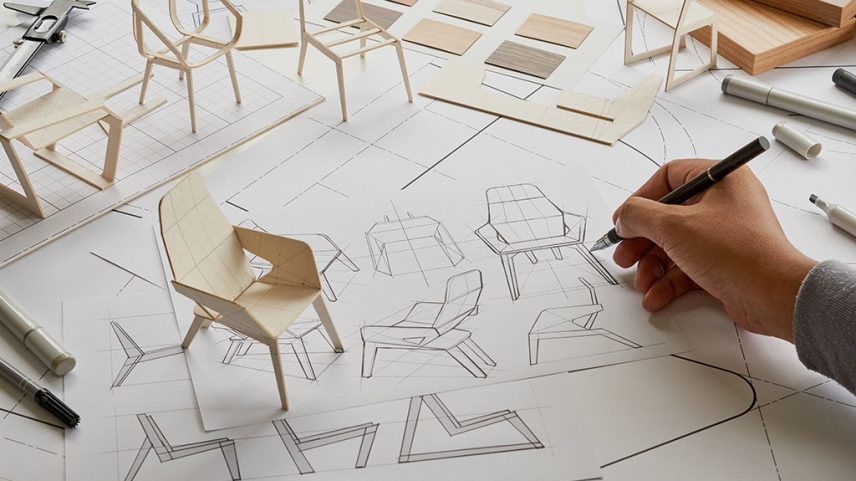 Mão desenhando esboços de cadeiras em papel quadriculado, com modelos em miniatura de madeira e amostras de materiais ao redor