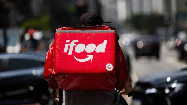A imagem mostra um entregador de bicicleta visto de costas, usando uma mochila vermelha com o logotipo do iFood. O fundo apresenta uma rua movimentada com carros e edifícios ao longe.