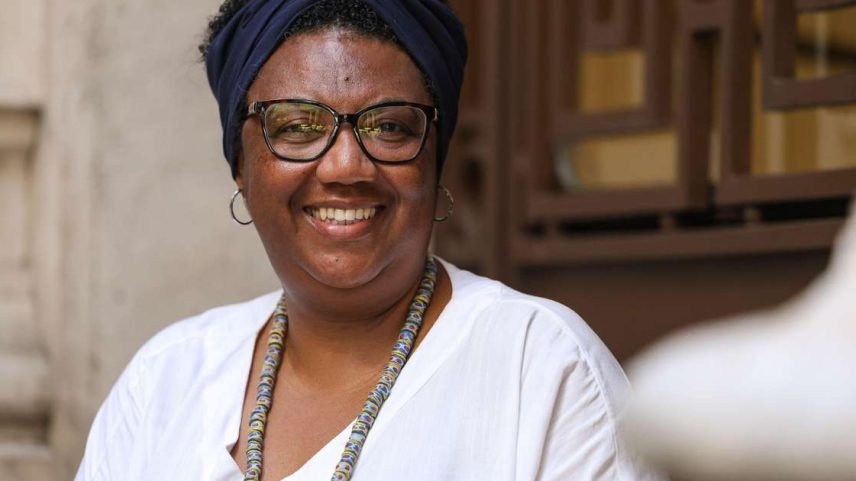 Rosana Paulino é uma mulher negra. Ela usa óculos de grau com armação preta, uma faixa preta no cabelo curto e uma blusa branca com um colar.