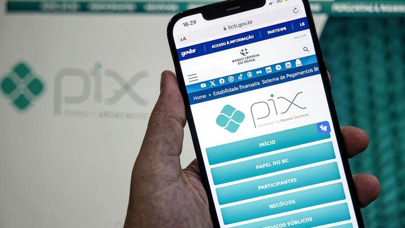 A imagem mostra uma mão segurando um smartphone que exibe a interface do aplicativo Pix. Na tela do celular, é possível ver o logotipo do Pix e opções como 'INÍCIO', 'PAINEL BC', 'PARTICIPANTES', 'NEGÓCIOS', 'SERVIÇOS PÚBLICOS' e 'ACESSIBILIDADE'. Ao fundo, há uma tela com o logotipo do Pix em um tom mais claro. O ambiente parece ser um local de trabalho ou um escritório.
