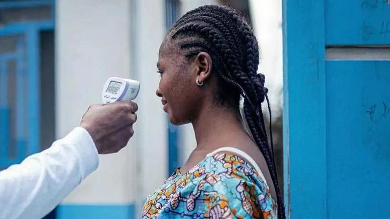 Um médico verifica a temperatura de uma mulher usando um termômetro infravermelho sem contato. Somente o braço do médico, que está vestido de branco, é mostrado. A mulher que está com a temperatura verificada tem o cabelo trançado. Ela está vestindo uma blusa azul brilhante com um padrão e está ao lado de uma porta de madeira azul com um prédio branco e azul ao fundo.
