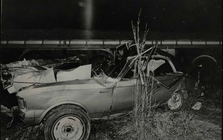 A imagem em preto e branco mostra um carro conversível gravemente danificado após um acidente. O veículo parece estar destruído, com a parte traseira completamente esmagada e a estrutura deformada. O local do acidente parece ser uma área rural ou uma estrada com vegetação ao redor. No fundo, há uma estrutura que pode ser um viaduto ou uma ponte. Um homem, possivelmente um policial ou investigador, observa a cena no interior do carro. A atmosfera da foto sugere um evento trágico ocorrido à noite