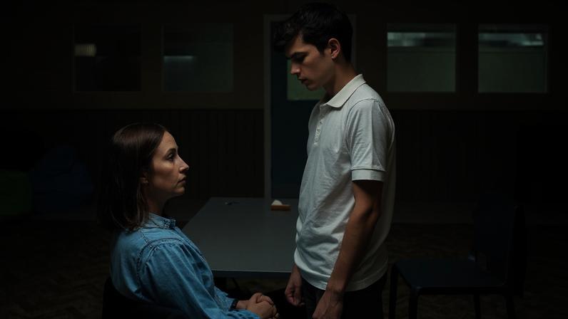 A imagem mostra um ambiente interno com duas pessoas em foco. Uma mulher está sentada em uma cadeira, olhando para um jovem que está em pé à sua frente, segurando suas mãos. O ambiente é simples, com paredes claras e algumas janelas ao fundo. Há também cadeiras e pufes coloridos visíveis.