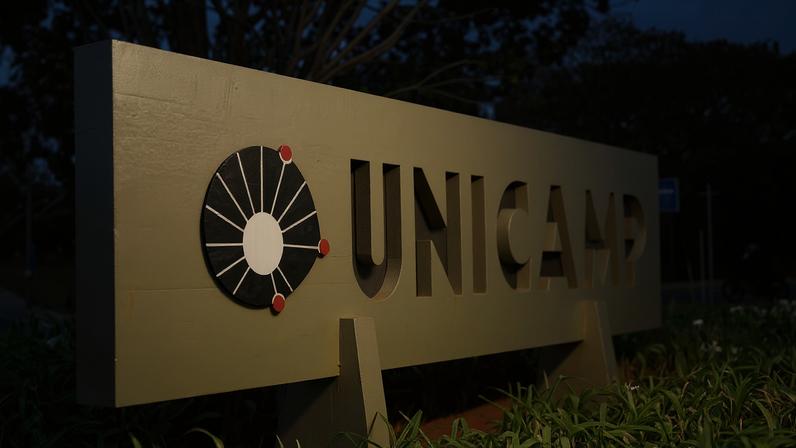 A imagem mostra uma placa de sinalização da Universidade Estadual de Campinas (UNICAMP). A placa é de cor verde e apresenta o nome 'UNICAMP' em letras grandes e em destaque. À esquerda do texto, há um símbolo gráfico que parece representar um círculo com linhas e pontos. Ao fundo, é possível ver árvores e um caminho, sugerindo um ambiente ao ar livre.