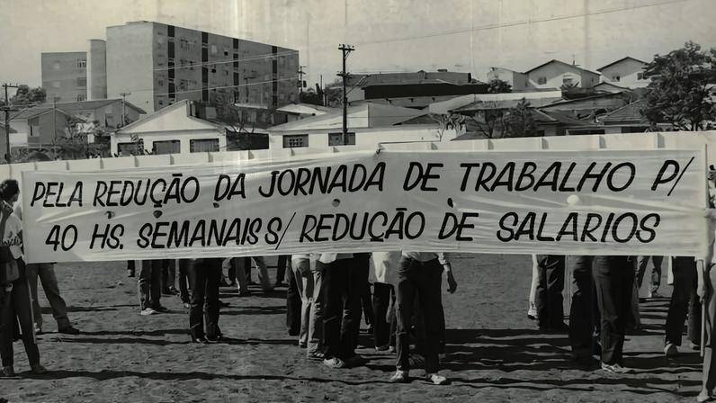 A imagem mostra um grupo de pessoas em um protesto, segurando um grande banner que diz: 'PELA REDUÇÃO DA JORNADA DE TRABALHO P/ 40 HS. SEMANAIS / REDUÇÃO DE SALÁRIOS'. O cenário é um campo aberto, com algumas edificações ao fundo. As pessoas estão vestidas de forma casual e parecem estar organizadas em uma manifestação.