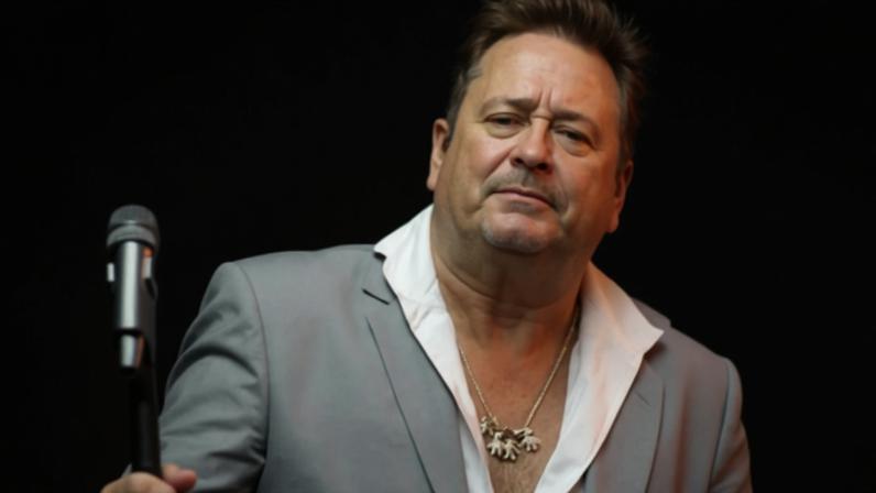 Um cantor em uma apresentação ao vivo, usando um terno cinza e uma camisa branca. Ele está segurando um microfone e parece estar cantando com uma expressão intensa. O fundo é desfocado, mas sugere um ambiente de show com luzes e uma plateia.