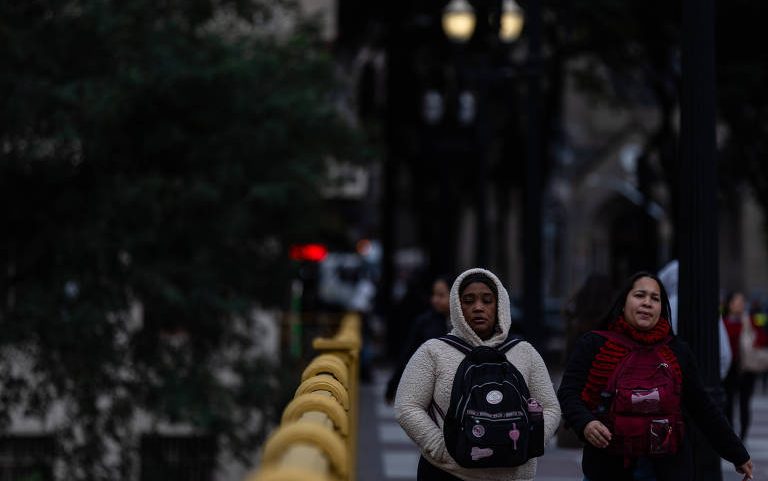 Duas mulheres caminham em uma rua, uma delas usa um moletom claro e a outra um casaco vermelho. Ao fundo, há árvores e postes de luz, com uma iluminação suave. A cena parece ser ao entardecer, com um ambiente urbano.