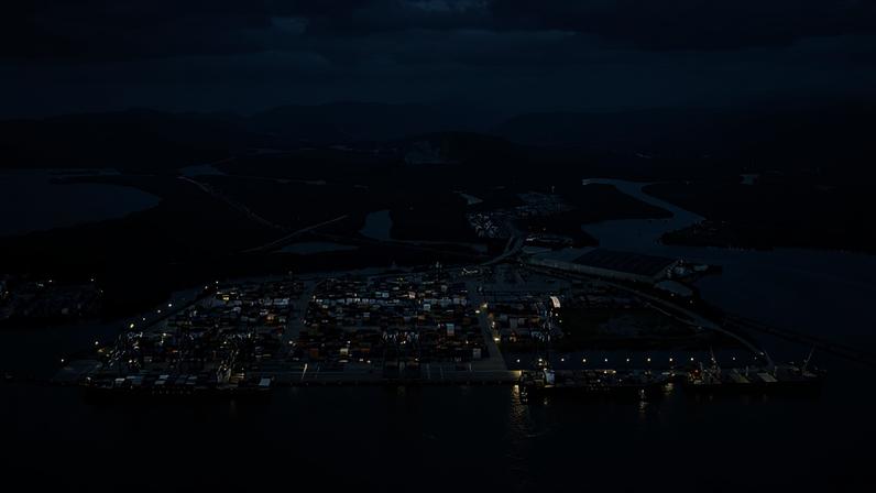 Vista aérea de um porto com contêineres e navios atracados. O cenário inclui áreas de água, vegetação ao redor e montanhas ao fundo, com um céu nublado.