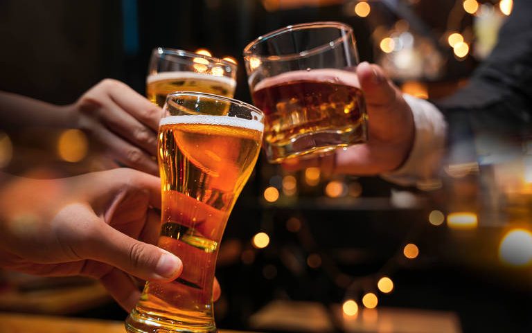 A imagem mostra várias mãos segurando copos de cerveja em um brinde. Os copos são de diferentes formatos, com uma cerveja clara e espumosa. O fundo é desfocado, com luzes brilhantes que criam um ambiente festivo.