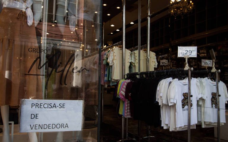 A imagem mostra a entrada de uma loja de roupas, onde há um cartaz com a inscrição 'Precisa-se de vendedora'. Ao fundo, são visíveis várias peças de roupas penduradas em cabides, incluindo camisetas de diferentes cores.