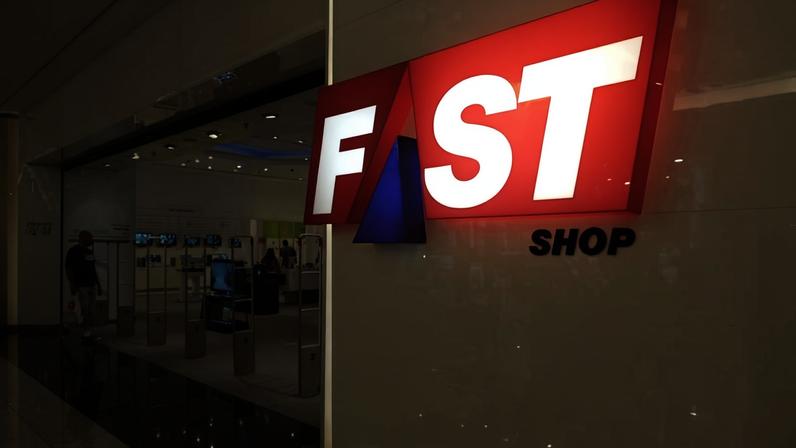 A imagem mostra a entrada de uma loja chamada Fast Shop, com um grande letreiro em destaque. O letreiro é composto pelas palavras 'FAST' em letras grandes e vermelhas, e 'SHOP' em letras menores e azuis. Ao fundo, é possível ver o interior da loja, que possui expositores de produtos eletrônicos e algumas pessoas dentro. O piso é de mármore e reflete a luz.
