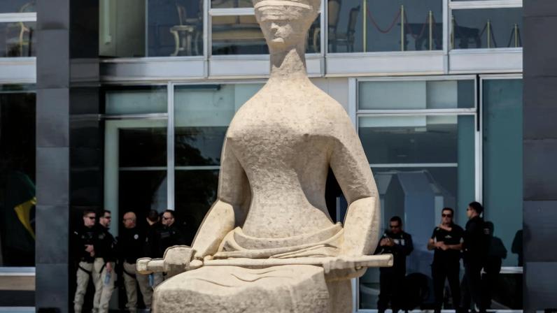 A imagem mostra uma escultura da Justiça em primeiro plano, com uma figura feminina de olhos vendados e segurando uma balança. Ao fundo, há um grupo de policiais em trajes táticos, observando a área. O ambiente parece ser um espaço público, com grandes janelas e uma decoração moderna.