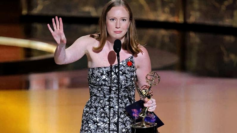 Uma mulher em um vestido preto e branco está em pé em um palco, segurando um prêmio Emmy em uma mão e um papel na outra. Ela está falando ao microfone e gesticulando com a mão livre. O fundo é iluminado com luzes quentes, e o ambiente parece ser uma cerimônia de premiação.