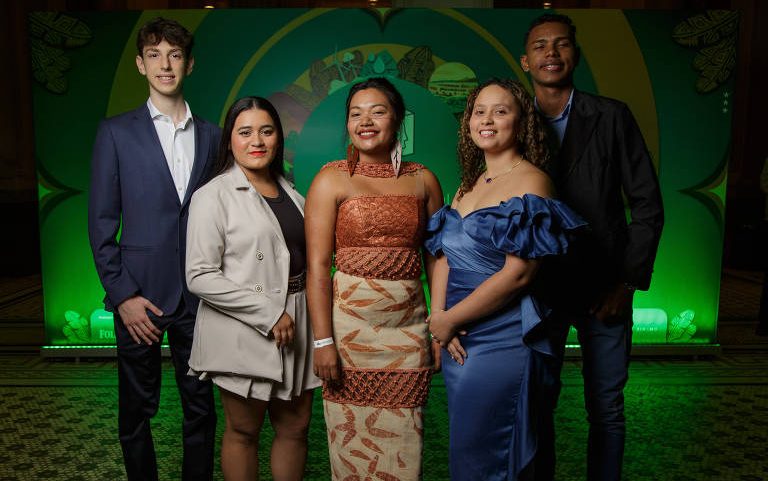 Cinco jovens posam lado a lado em ambiente interno com iluminação verde ao fundo. Dois homens vestem ternos escuros, três mulheres usam vestidos elegantes em tons bege, marrom e azul. Piso tem padrão geométrico.