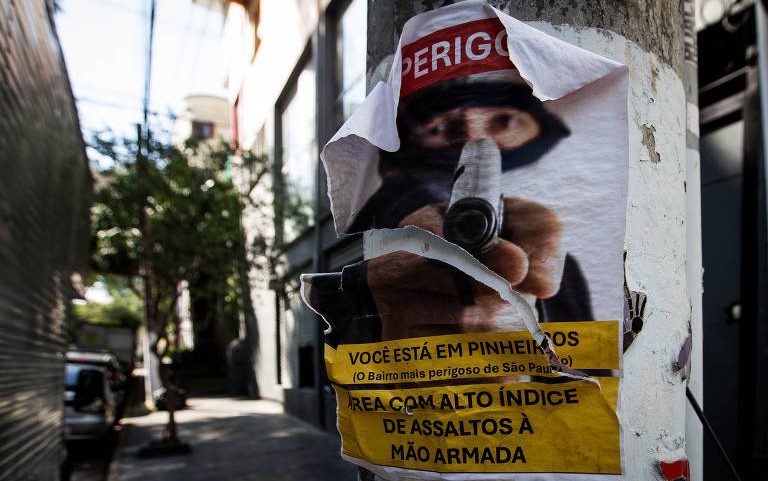 Cartaz rasgado colado em poste na rua alerta para perigo em Pinheiros, São Paulo, destacando alto índice de assaltos à mão armada. Imagem mostra figura com arma apontada e texto em amarelo e vermelho.