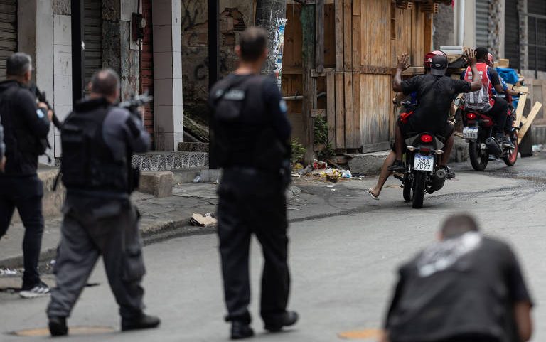 Quatro policiais armados observam dois motociclistas em rua estreita com construções simples e grafites nas paredes. Um policial está agachado na calçada, enquanto os motociclistas avançam pela via.