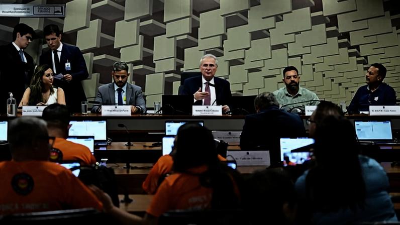 Cinco autoridades sentadas em mesa de plenário discutem, com público em cadeiras à frente, alguns usando camisetas laranja. Dois homens em pé conversam ao fundo. Ambiente interno com teto decorado por painéis geométricos.